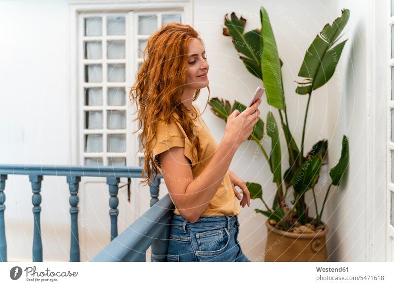 Lächelnde rothaarige junge Frau lehnt am Geländer und schaut auf Handy Informationen T-Shirts Telekommunikation Handies Handys Mobiltelefon Mobiltelefone