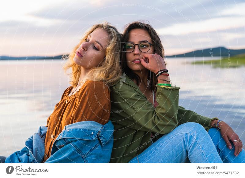 Finnland, Lappland, zwei junge Frauen sitzen Rücken an Rücken am Seeufer in der Dämmerung Seen Daemmerung weiblich Freundin sitzend sitzt Gewässer Wasser