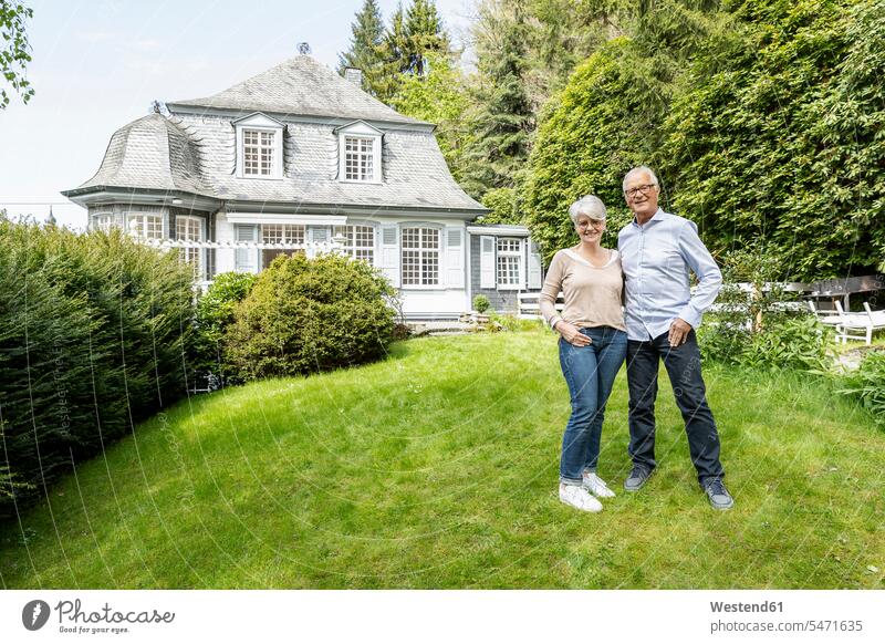 Lächelndes älteres Ehepaar steht im Garten seines Hauses Brillen freuen geniessen Genuss Glück glücklich sein glücklichsein zufrieden stehend daheim zu Hause