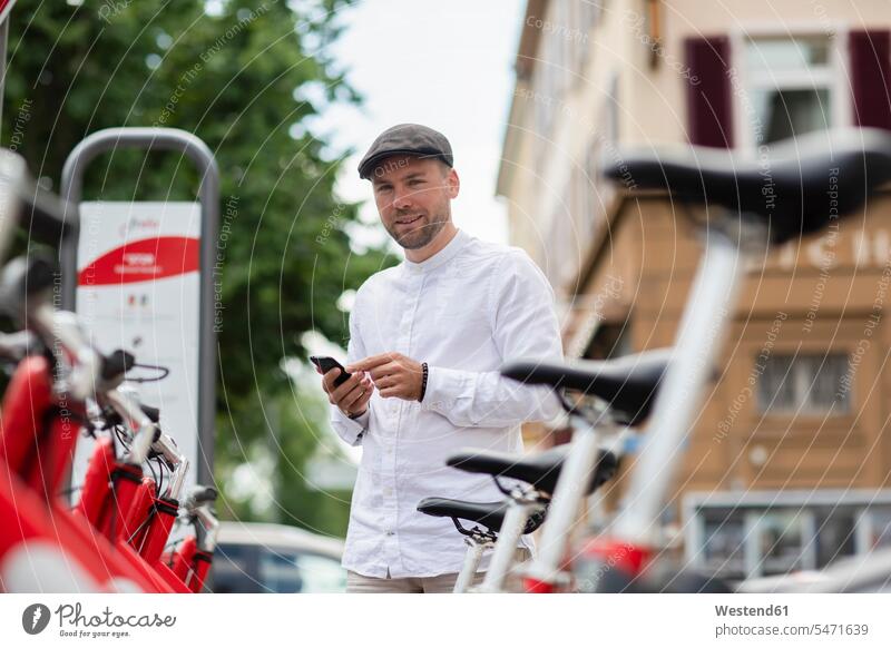 Lächelnder Mann mietet Fahrrad über Smartphone am Parkplatz in der Stadt Farbaufnahme Farbe Farbfoto Farbphoto Deutschland Außenaufnahme außen draußen im Freien