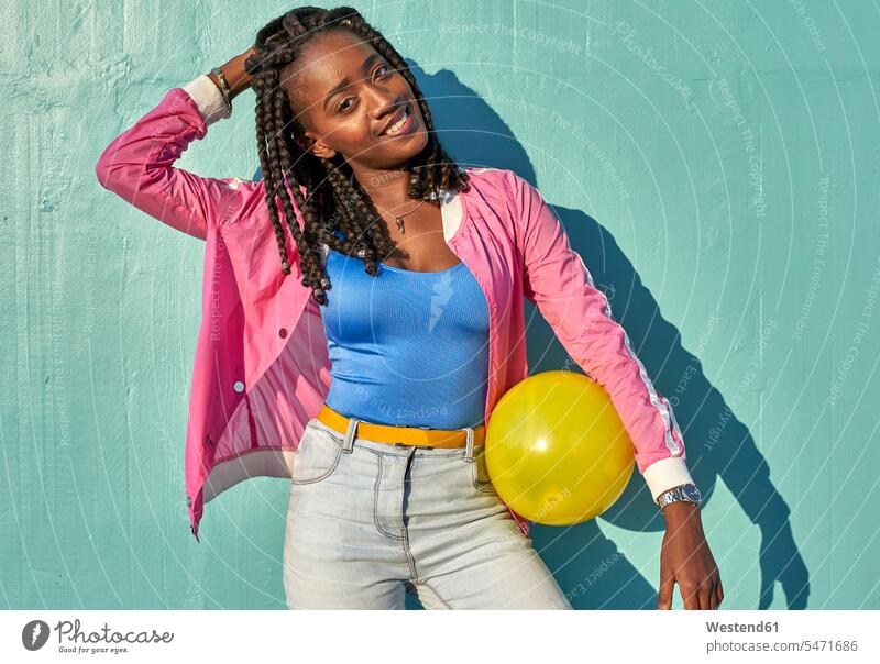 Junge schwarze Frau mit einem gelben Ball vor einer blauen Wand Jacken Bälle freuen Glück glücklich sein glücklichsein gefühlvoll Emotionen Empfindung