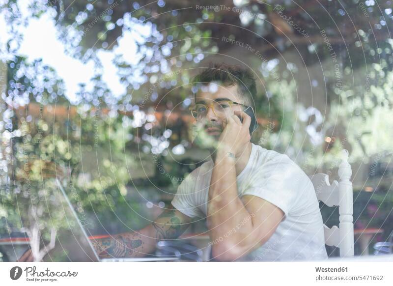 Junger Mann mit Laptop am Handy hinter der Fensterscheibe eines Cafés Fensterscheiben T-Shirts Rechner Laptops Notebook Notebooks Telekommunikation telefonieren