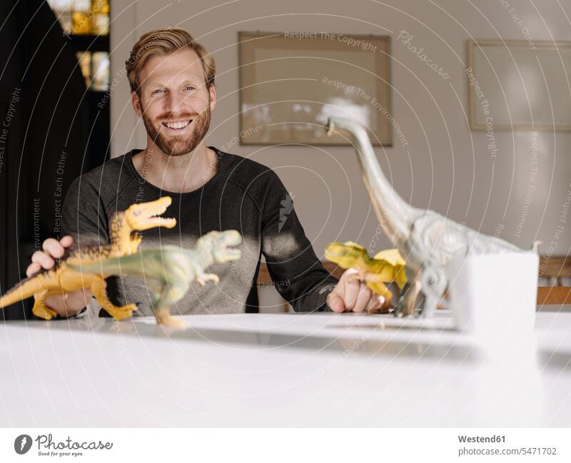 Porträt eines lächelnden Mannes mit Spielzeugdinosauriern auf dem Tisch Leute Menschen People Person Personen Europäisch Kaukasier kaukasisch 1 Ein ein Mensch