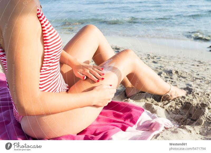 Frau sitzt am Strand und trägt Sonnencreme auf Handtuecher Handtücher Badebekleidung Badeanzuege Badeanzüge Cremes Sonnenmilch cremen entspannen relaxen sitzend