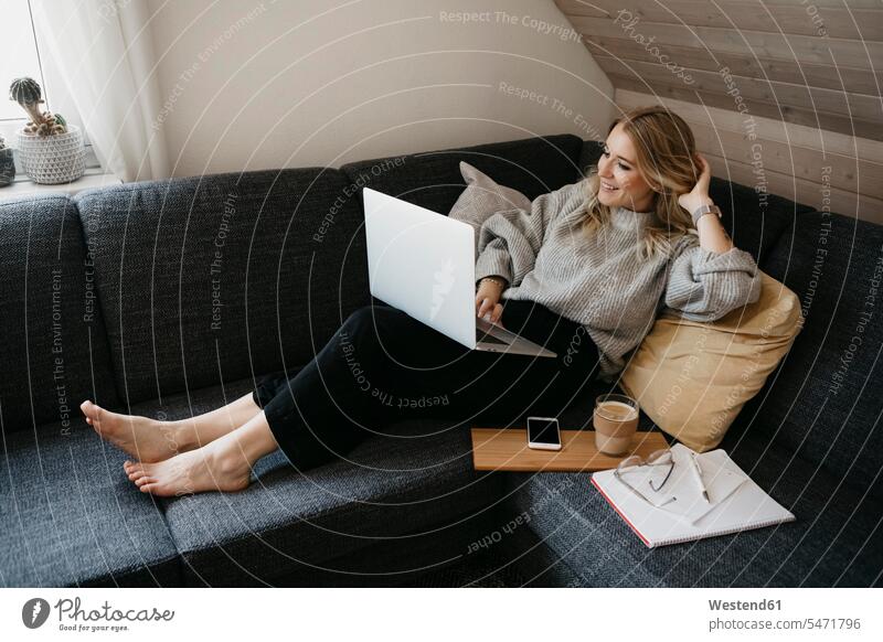 Lächelnde Frau benutzt Laptop auf Sofa, während sie zu Hause freiberuflich arbeitet Farbaufnahme Farbe Farbfoto Farbphoto Innenaufnahme Innenaufnahmen innen