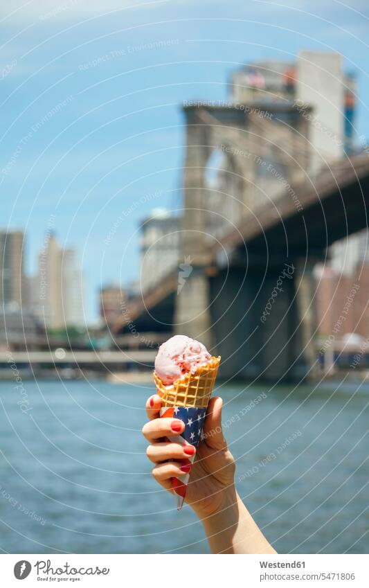 USA, New York, Brooklyn, Nahaufnahme der Hand einer Frau, die eine Erdbeereistüte mit der Brooklyn Bridge im Hintergrund hält Eistüte Eistuete Eiswaffel