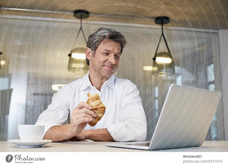 Geschäftsmann mit Laptop beim Croissant-Essen Notebook Laptops Notebooks essen essend Hoernchen Croissants Cornetto Cornettos Hörnchen Businessmann