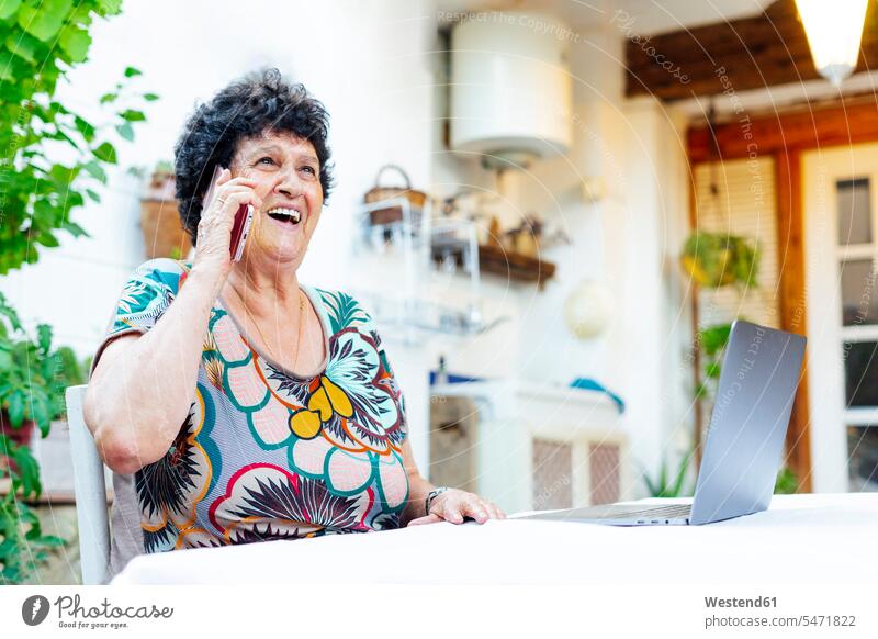 Fröhliche ältere Frau spricht über Smartphone, während sie im Hof am Laptop sitzt Farbaufnahme Farbe Farbfoto Farbphoto Spanien Freizeitbeschäftigung Muße Zeit