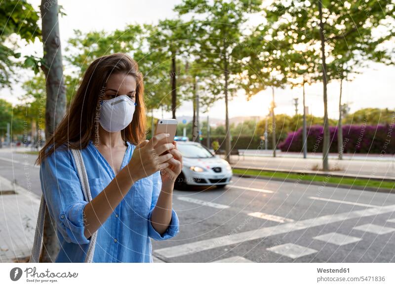 Junge Frau mit Maske, die ein Smartphone benutzt, während sie in der Stadt auf der Straße steht Farbaufnahme Farbe Farbfoto Farbphoto Spanien Freizeitkleidung