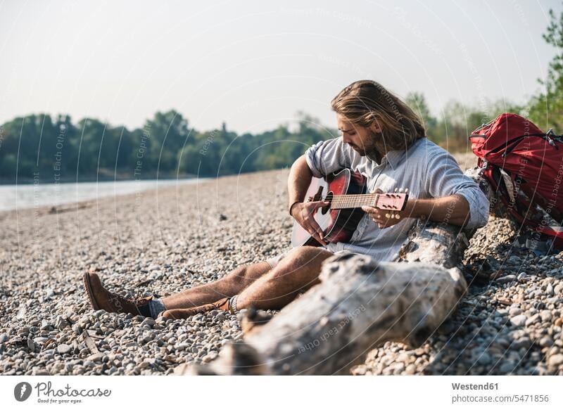 Junger Mann sitzt am Flussufer und spielt Gitarre Fluesse Fluß Flüsse Männer männlich spielen sitzen sitzend Gitarren Gewässer Wasser Ufer Erwachsener erwachsen