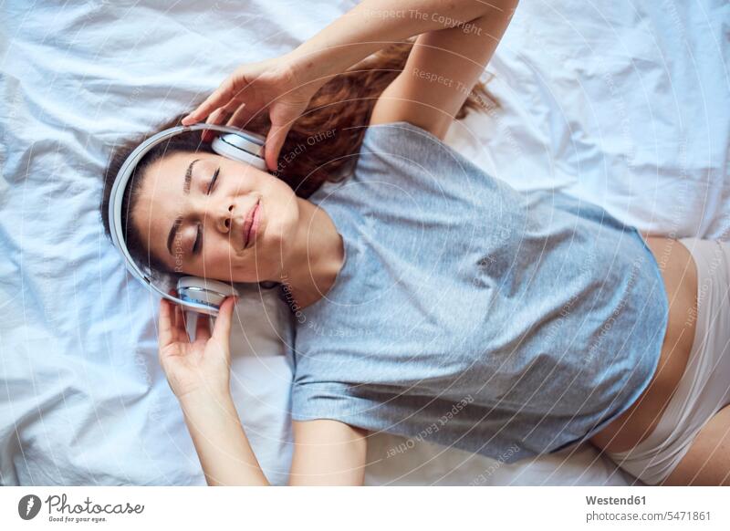 Porträt einer jungen Frau mit geschlossenen Augen auf dem Bett liegend und mit Kopfhörern Musik hörend Leute Menschen People Person Personen Europäisch