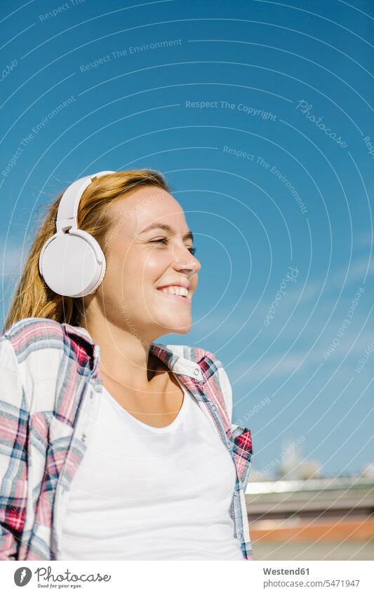 Glückliche Frau hört Musik über Kopfhörer und schaut an einem sonnigen Tag gegen den blauen Himmel Farbaufnahme Farbe Farbfoto Farbphoto Außenaufnahme außen