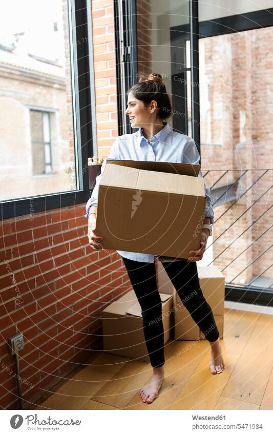 Lächelnde junge Frau trägt Karton in neuer Wohnung und schaut aus dem Fenster Pappkartons Kartons tragen transportieren weiblich Frauen Mietwohnungen wohnen