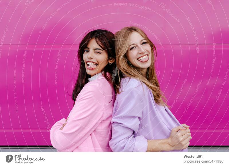 Schwestern strecken ihre Zunge heraus, während sie Rücken an Rücken an einer rosa Wand stehen Farbaufnahme Farbe Farbfoto Farbphoto Außenaufnahme außen draußen