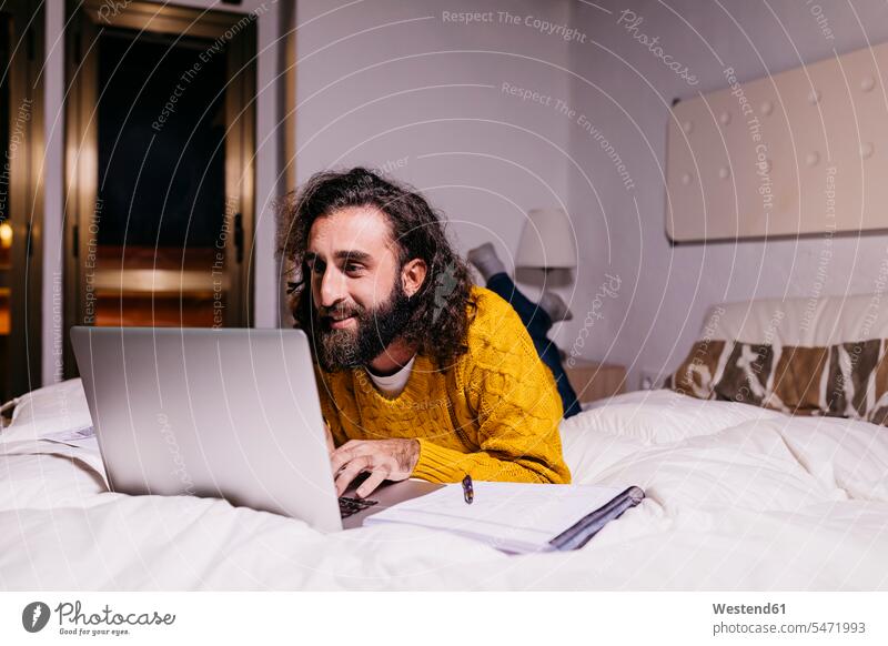 Lächelnder junger Mann liegt im Bett zu Hause mit Laptop Betten Männer männlich Zuhause daheim lächeln Notebook Laptops Notebooks liegen liegend Erwachsener