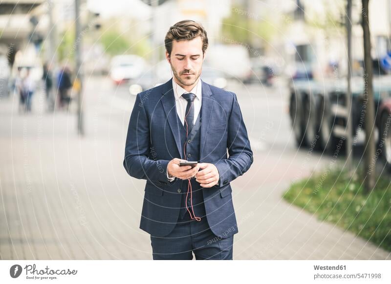 Geschäftsmann zu Fuß in der Stadt mit Handy Deutschland Ohrhörer Smartphone iPhone Smartphones Freiberufler freiberuflich freie Berufe Pendler Führungskraft