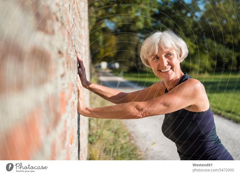 Lächelnde sportliche ältere Frau lehnt an einer Backsteinmauer weiblich Frauen anlehnen angelehnt lehnend lächeln Seniorin Seniorinnen alt Sport Erwachsener