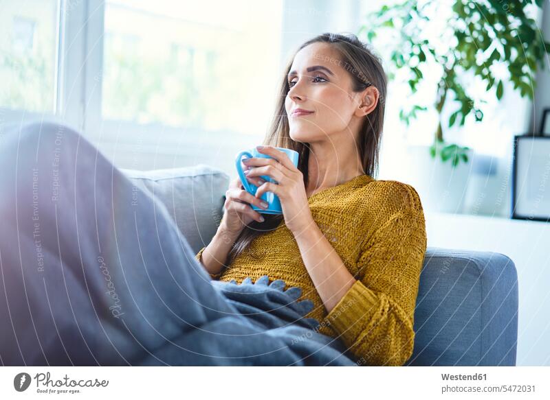 Hübsche junge Frau genießt eine Tasse Kaffee, während sie auf dem Sofa unter einer Decke liegt Leute Menschen People Person Personen Alleinstehende