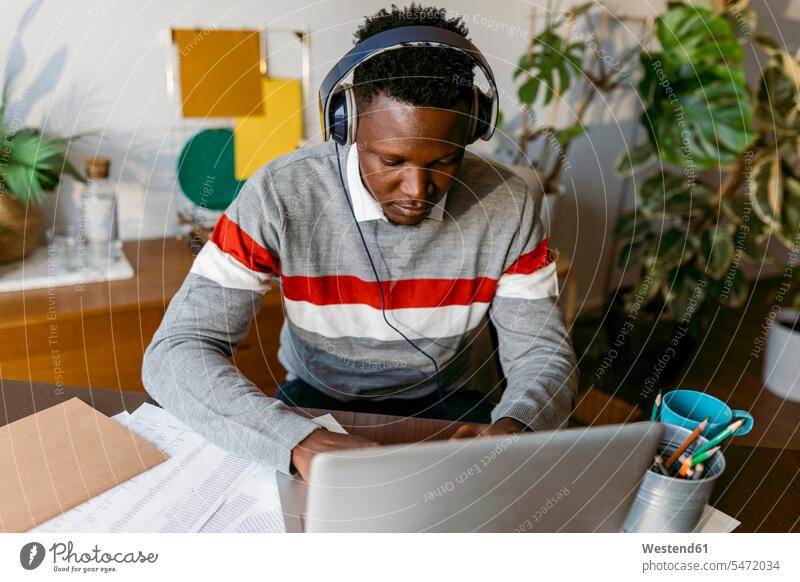 Afrikanischer Geschäftsmann mit Kopfhörern bei Videogespräch am Laptop zu Hause Farbaufnahme Farbe Farbfoto Farbphoto Innenaufnahme Innenaufnahmen innen drinnen