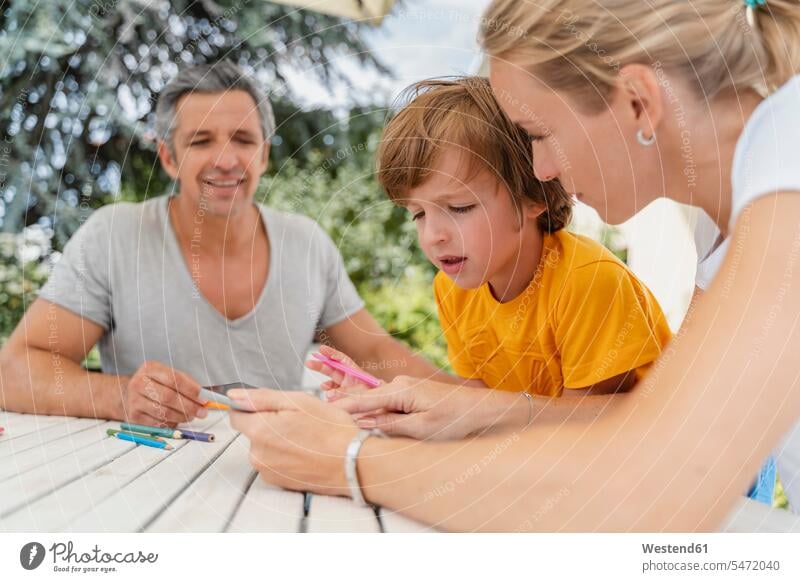 Vater, Mutter und Sohn machen gemeinsam Hausaufgaben auf der Terrasse Schüler T-Shirt Tisch sitzen Sommer Geborgenheit Zuhause Gemeinsam Harmonie Internet