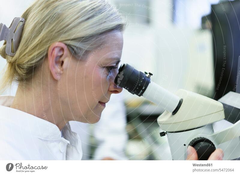Blonde Wissenschaftlerin schaut durch ein Mikroskop in ein beleuchtetes Labor Farbaufnahme Farbe Farbfoto Farbphoto Innenaufnahme Innenaufnahmen innen drinnen