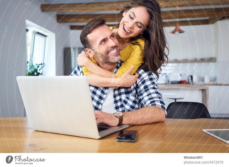 Glückliches Paar, das am Esstisch sitzt, sich umarmt und einen Laptop benutzt Laptop benutzen Laptop benützen glücklich glücklich sein glücklichsein Tisch