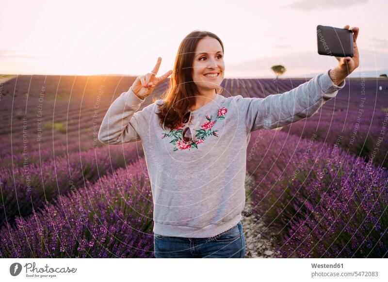 Frankreich, Valensole, Porträt einer lächelnden Frau, die ein Selfie vor einem Lavendelfeld bei Sonnenuntergang macht Portrait Porträts Portraits weiblich