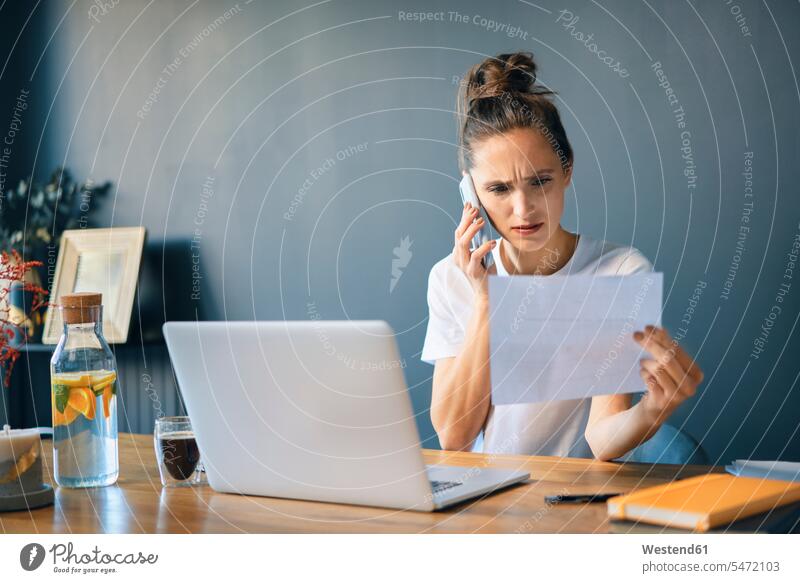 Geschäftsfrau hält Dokument in der Hand und spricht über Smartphone, während sie im Heimbüro am Schreibtisch sitzt Farbaufnahme Farbe Farbfoto Farbphoto