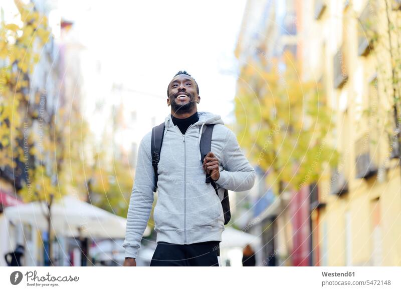Junger Mann mit Rucksack lächelnd beim Spaziergang in der Stadt Farbaufnahme Farbe Farbfoto Farbphoto Außenaufnahme außen draußen im Freien Tag
