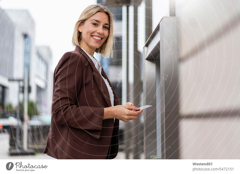 Porträt einer lächelnden jungen Geschäftsfrau, die an einem Geldautomaten in der Stadt Geld abhebt geschäftlich Geschäftsleben Geschäftswelt Geschäftsperson