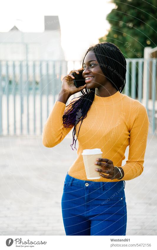 Glückliche Teenager-Mädchen mit Kaffeetasse Blick weg während des Gesprächs auf Smartphone auf der Straße Farbaufnahme Farbe Farbfoto Farbphoto Außenaufnahme