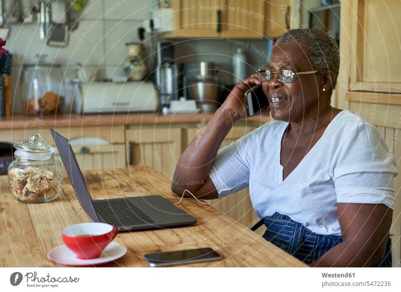Ältere Frau sitzt am Küchentisch mit Laptop und Smartphone Leute Menschen People Person Personen Afrikanisch Afrikanische Abstammung dunkelhäutig Farbige