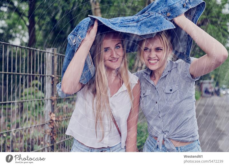 Zwei junge Frauen, die mit einer Jeansjacke vor dem Regen schlottern Freundinnen Jeansjacken weiblich Hecke Hecken beschützen Freunde Freundschaft Kameradschaft