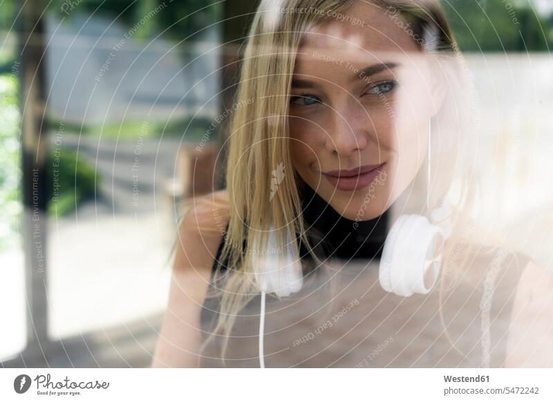 Porträt einer lächelnden blonden Frau mit Kopfhörern hinter einer Fensterscheibe Fensterscheiben Lifestyle Lebensstil Schönheit Beauty Zuversicht Zuversichtlich