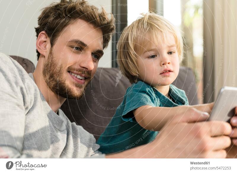 Vater und Sohn schauen sich zu Hause auf der Couch ein Smartphone an Sofa Couches Liege Sofas ansehen Zuhause daheim Papas Väter Vati Vatis Papis iPhone