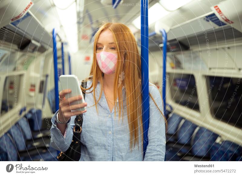 Junge Frau mit Maske, die im Zug stehend ein Smartphone benutzt Farbaufnahme Farbe Farbfoto Farbphoto Zuginnenraum Bahn Züge Bahnen Verkehrswesen Transportwesen