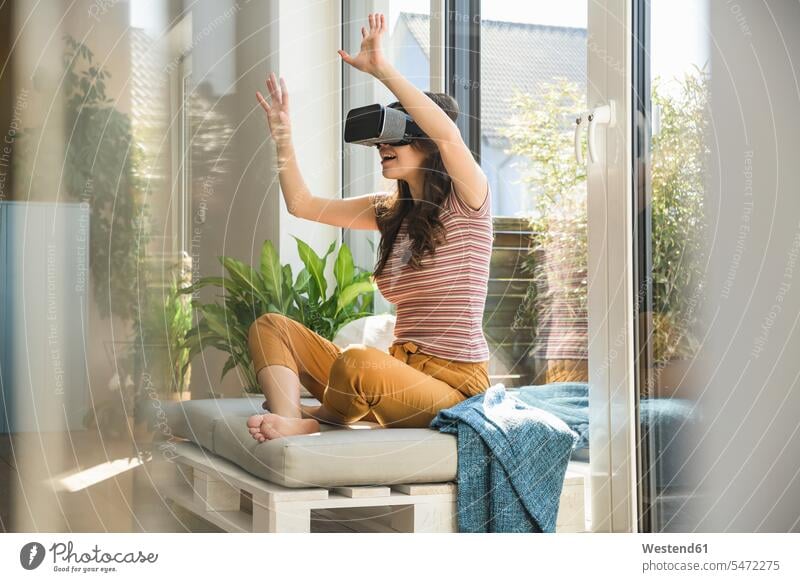 Junge Frau sitzt am Fenster zu Hause mit VR-Brille weiblich Frauen virtuell Virtualität Brillen Zuhause daheim sitzen sitzend Erwachsener erwachsen Mensch