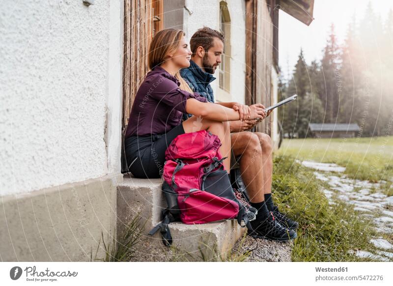 Junges Paar mit Karte auf einem Bauernhof während einer Wanderung, Vorderriss, Bayern, Deutschland Touristen Rucksäcke Karten Landkarten Hemden sitzend sitzt