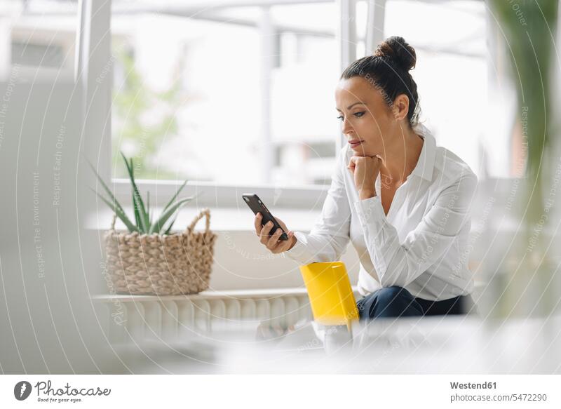 Unternehmerin benutzt Mobiltelefon, während sie im Home-Office am Fenster sitzt Farbaufnahme Farbe Farbfoto Farbphoto Deutschland Innenaufnahme Innenaufnahmen