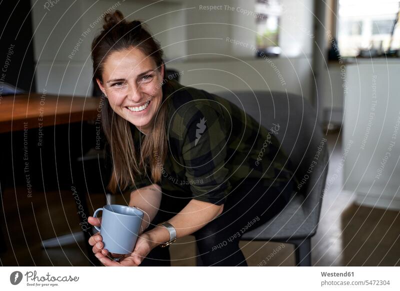 Lächelnde Frau hält Kaffeetasse und schaut weg, während sie zu Hause auf einem Stuhl sitzt Farbaufnahme Farbe Farbfoto Farbphoto Deutschland
