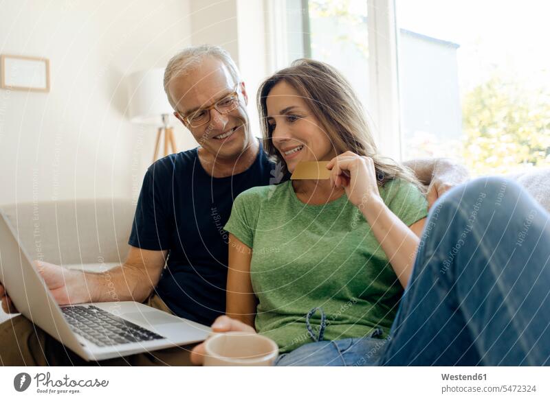 Lächelnd reifen Paar sitzt auf der Couch zu Hause einkaufen online mit Laptop Couches Liege Sofas Rechner Laptops Notebook Notebooks sitzend daheim Sharing