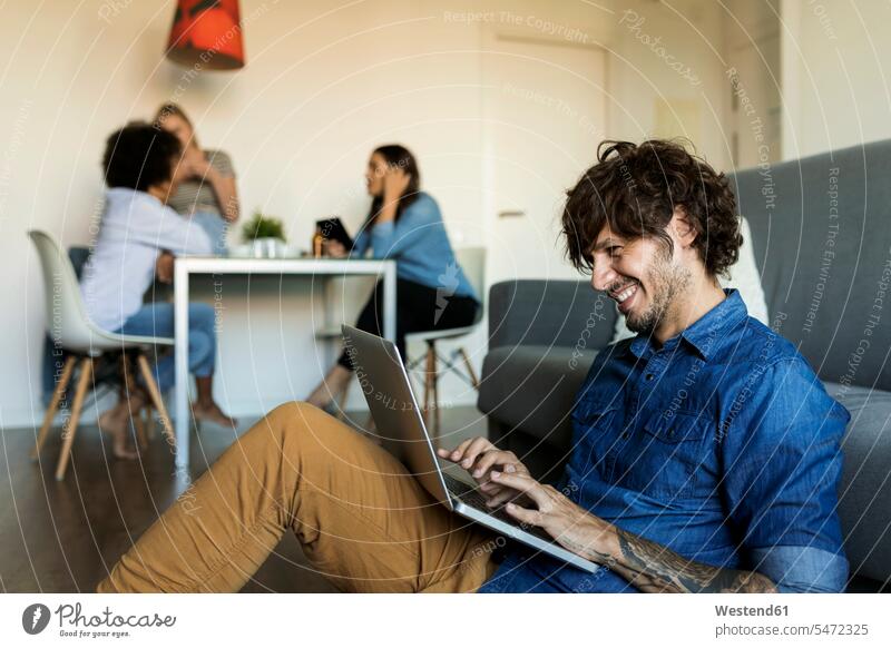 Lächelnder Mann sitzt auf dem Boden mit Laptop mit Freunden im Hintergrund Böden Boeden Notebook Laptops Notebooks Männer männlich sitzen sitzend lächeln