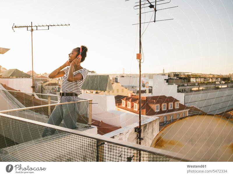 Glückliche junge Frau steht bei Sonnenuntergang auf dem Dach und hört Musik mit Kopfhörern Leute Menschen People Person Personen Afrikanisch