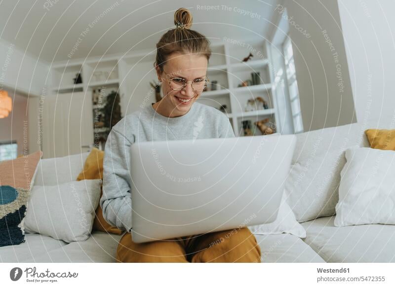 Lächelnde Frau arbeitet am Laptop, während sie zu Hause auf dem Sofa sitzt Farbaufnahme Farbe Farbfoto Farbphoto Innenaufnahme Innenaufnahmen innen drinnen Tag