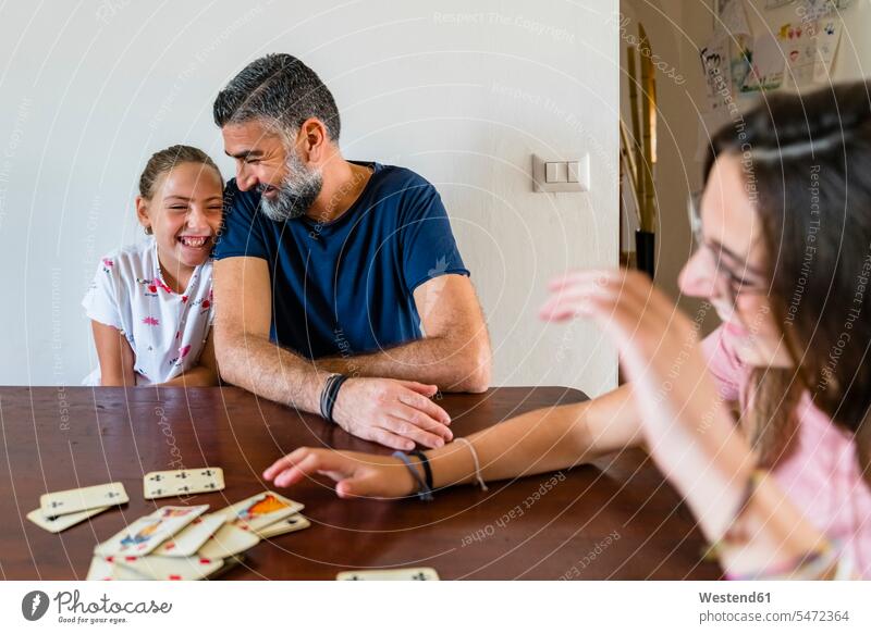 Glücklicher Vater mit zwei Töchtern, die zu Hause auf einem Holztisch Karten spielen Spielkarten T-Shirts Tische Holztische Spiele Gesellschaftsspiele