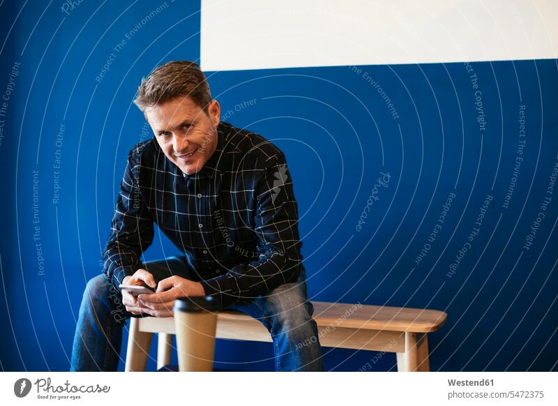 Porträt eines lächelnden Geschäftsmannes, der auf einer Bank sitzt und ein Mobiltelefon hält Businessmann Businessmänner Geschäftsmänner Portrait Porträts