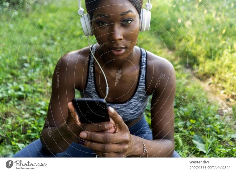Junger Sportler in der Natur, hört Musik mit Kopfhörern, hält Smartphone iPhone Smartphones Joggerin Joggerinnen hören hoeren Hocke hocken Sommer Sommerzeit