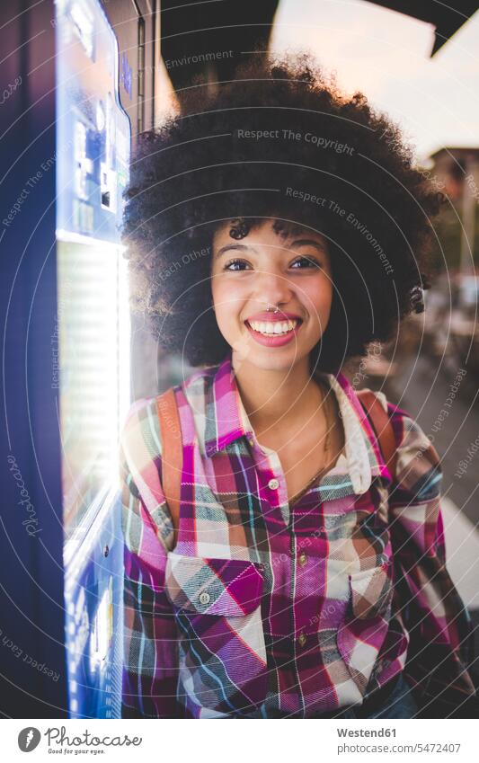 Porträt einer glücklichen jungen Frau mit Afro-Frisur, die in der Abenddämmerung in der Stadt an einen Automaten gelehnt ist Leute Menschen People Person