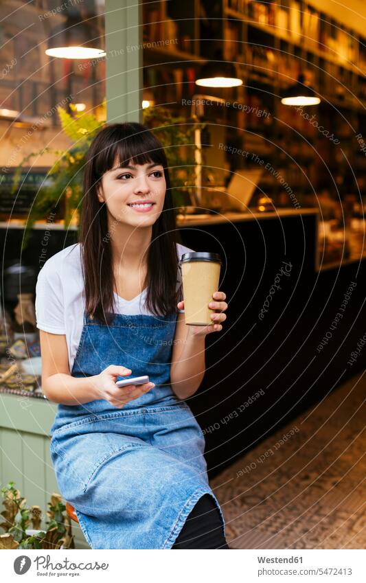 Lächelnde Frau sitzt an der Eingangstür eines Geschäfts mit Handy und Kaffee zum Mitnehmen sitzen sitzend lächeln Shop Laden Läden Geschäfte Shops weiblich