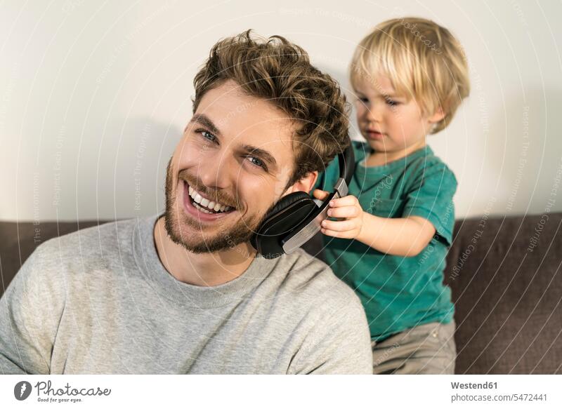 Glückliche Vater und Sohn mit Kopfhörern auf der Couch zu Hause glücklich glücklich sein glücklichsein Papas Väter Vati Vatis Papis aufsetzen Zuhause daheim